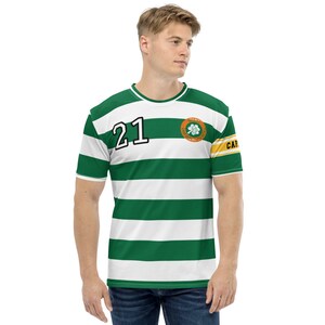 Pode incluir: Uma camiseta listrada verde e branca com um logotipo de trevo verde e o número 21 na frente. A camiseta tem uma faixa amarela e laranja na manga direita com a palavra "CAPTAIN" impressa nela.