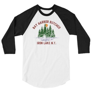 May include: A white and black baseball style t-shirt with a vintage graphic design. The graphic features a sunset over a lake with trees and the text "Bay Harbor Butcher Iron Lake N.Y."