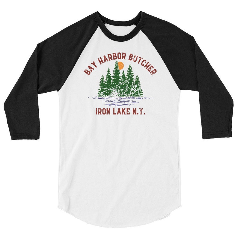 May include: A white and black baseball style t-shirt with a vintage graphic design. The graphic features a sunset over a lake with trees and the text "Bay Harbor Butcher Iron Lake N.Y."
