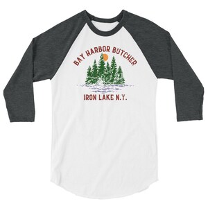May include: A white and grey baseball tee shirt with a distressed graphic design of a forest scene with a red sun and the text "Bay Harbor Butcher Iron Lake N.Y."