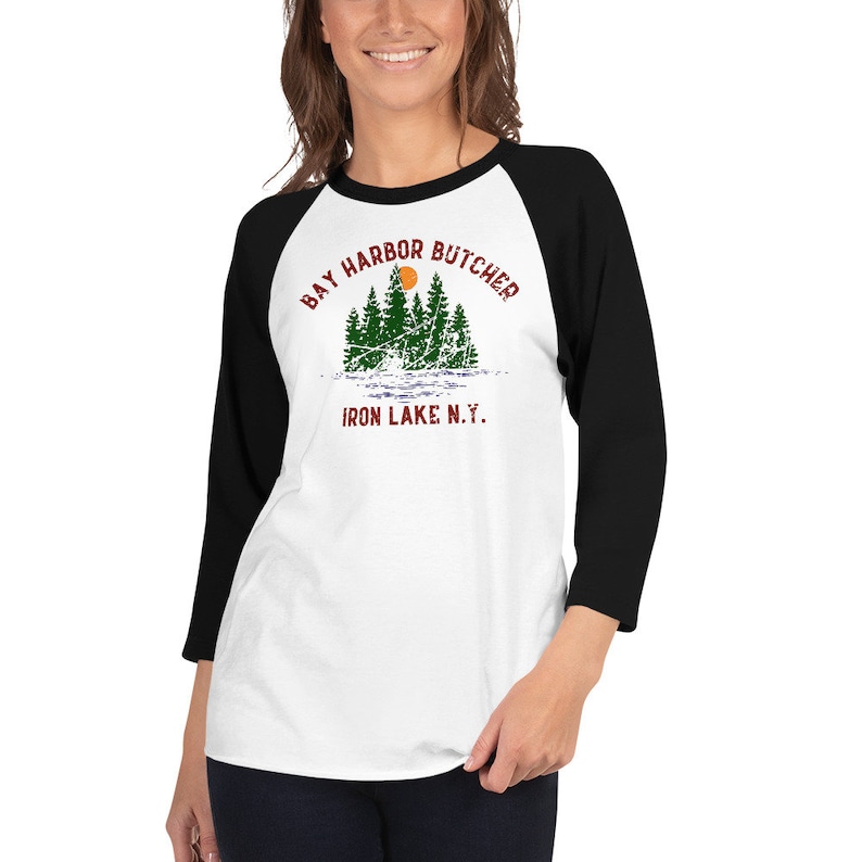 May include: A white and black baseball tee shirt with a vintage-style graphic of a sunset over a lake with trees. The text on the shirt reads "Bay Harbor Butcher Iron Lake N.Y."