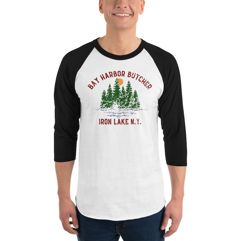 May include: A white and black baseball tee shirt with a distressed graphic design. The graphic features a sunset over a lake with trees and the text "Bay Harbor Butcher Iron Lake N.Y."