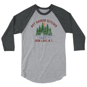 May include: A grey and black baseball tee shirt with a vintage-style graphic of a lake scene with trees and a sun. The text "Bay Harbor Butcher Iron Lake N.Y." is printed on the shirt.