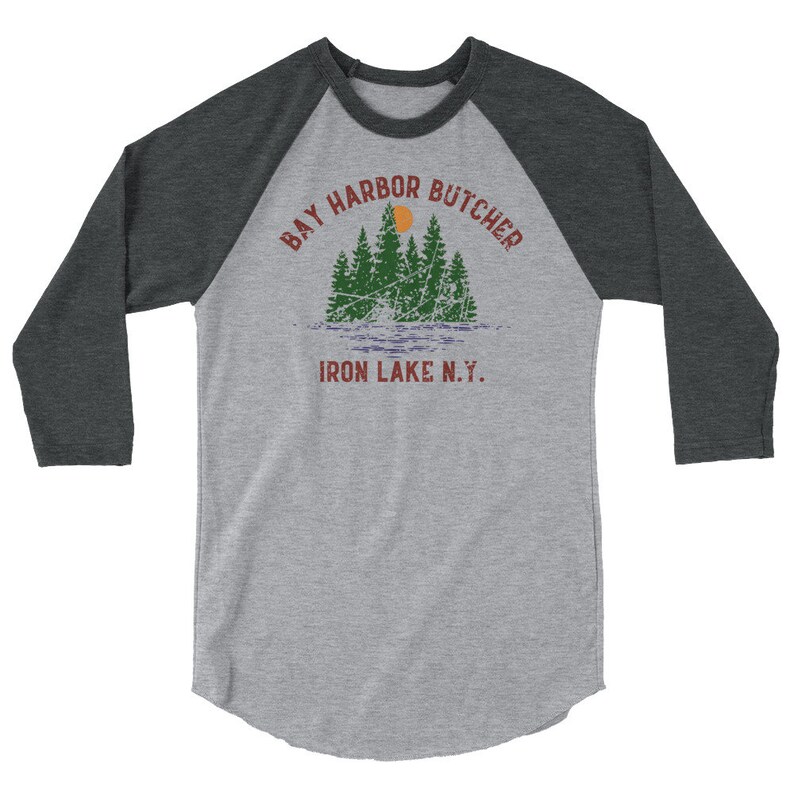 May include: A grey and black baseball tee shirt with a vintage-style graphic of a lake scene with trees and a sun. The text "Bay Harbor Butcher Iron Lake N.Y." is printed on the shirt.
