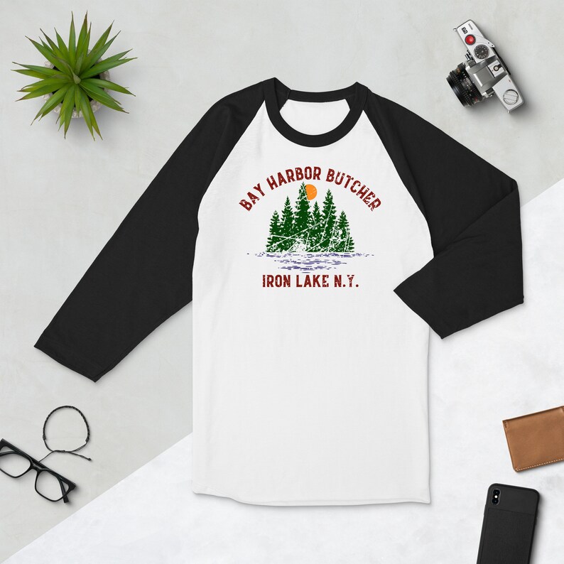 May include: A white and black baseball tee shirt with a red and green graphic design. The graphic features a group of trees with a sun and the text "Bay Harbor Butcher Iron Lake N.Y."