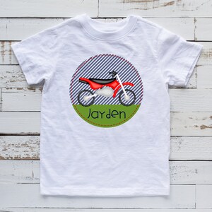 May include: White t-shirt with a red and black dirt bike graphic inside a blue and white striped circle with a green border. The text "Jayden" is written in black below the graphic.