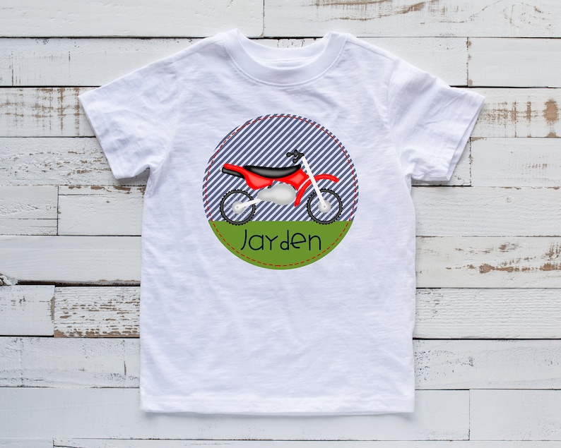 May include: White t-shirt with a red and black dirt bike graphic inside a blue and white striped circle with a green border. The text "Jayden" is written in black below the graphic.