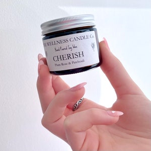 May include: A small, dark glass jar with a silver lid, labeled "CHERISH" with "Plum Rose & Patchouli" scent. The jar is held against a white background. The text "THE WELLNESS CANDLE CO." and "Hand Poured Soy Wax" are also visible.