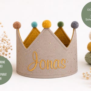 Könnte beinhalten: Eine beigefarbene Stoffkrone mit dem in Goldfaden gestickten Namen Jonas. Die Krone ist mit fünf kleinen Pompons in den Farben Braun, Türkis, Gelb, Grün und Grau verziert. Der Außenstoff weist ein dezentes, goldenes Sprenkelmuster auf.