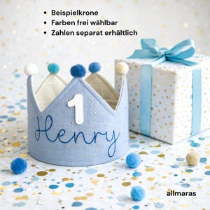 Könnte beinhalten: Eine hellblaue Stoffkrone mit dem Namen "Henry" in blauer Schrift und der Zahl "1" in Weiß. Die Krone ist mit blauen und cremefarbenen Pompons verziert. Im Hintergrund eine Geschenkbox mit blauem Band.