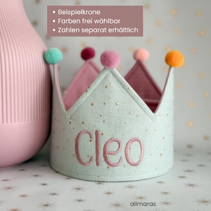Könnte beinhalten: Eine hellblaue Stoffkrone mit dem Namen "Cleo" in Rosa gestickt, verziert mit goldenen Sternen und mit bunten Pompons versehen. Der Text auf dem Bild lautet "Beispielkrone, Farben frei wählbar, Zahlen separat erhältlich."
