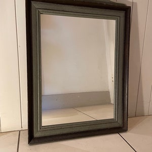May include: A vintage wooden mirror with a dark green painted frame and a decorative floral design painted on the top. The mirror has a simple rectangular shape and a bevelled edge.