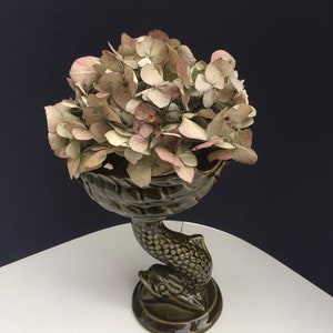 May include: A green ceramic vase shaped like a fish with a bowl on top, holding dried pink hydrangeas.