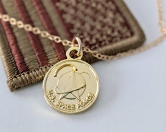 May include: A gold necklace with a round pendant featuring the U.S. Space Force logo and the words "U.S. Space Force". The necklace is lying on top of a brown and green embroidered patch with a design resembling the American flag.