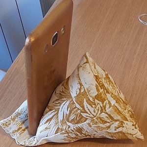 May include: A brown smartphone is propped up by a small, triangular pillow. The pillow is cream-colored with a repeating pattern of brown leaves and floral designs. The phone is angled slightly, resting against the pillow for support.