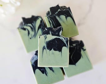 May include: A stack of four rectangular bars of soap featuring a marbled design with a light sage green base and deep black swirls. The soap bars have a smooth, solid texture with distinct, organic patterns created by the contrasting colors.