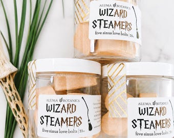 May include: Three clear plastic jars filled with light orange, bolt-shaped shower steamers. Each jar features a white label with the text ALUMA BOTANICS WIZARD STEAMERS five sinus love bolts 55oz and a graphic of a broomstick.