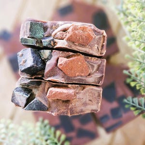 May include: Three bars of handmade soap stacked on top of each other. The soap is a dark brown color with embedded dark gray and reddish-brown stone-like pieces. The soap has a rustic, natural appearance.