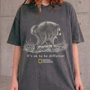 Pode incluir: Uma t-shirt cinzento-carvão de manga curta com uma ilustração de linhas brancas de um guaxinim na relva. Abaixo do gráfico, o texto diz "it's ok to be different" seguido pelo logótipo amarelo da National Geographic e o respetivo texto.