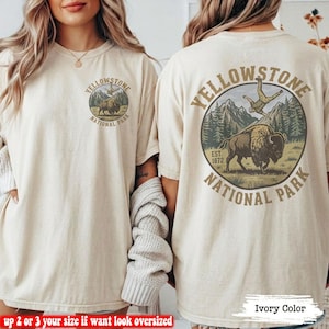May include: An ivory-colored t-shirt featuring a graphic of a bison and a person in a mountain landscape. The front has a small circular logo, while the back displays a larger version of the same design with the text Yellowstone National Park.