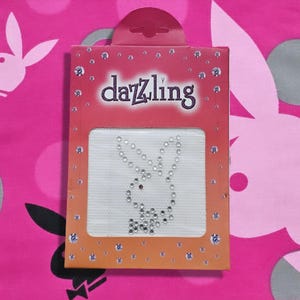 May include: A rectangular package with the word "dazzling" in purple lettering. The package contains a white patch with a bunny design made of rhinestones. The background is pink with a bunny logo.