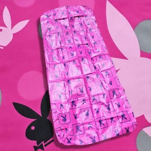 May include: A pink, rectangular item with a repeating pattern of the Playboy bunny logo in a square. The background is a pink fabric with a large bunny silhouette and polka dots. The item appears to be a pillow or blanket.