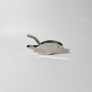 May include: A silver fish-shaped citrus juicer with a lever handle. The juicer is designed to squeeze lemons, limes, and oranges.