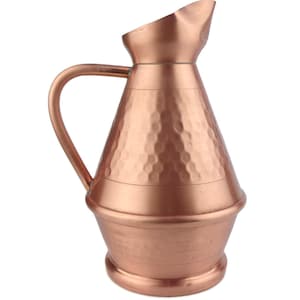 Puede incluir: Una jarra de cobre martillado con una base ancha y redondeada y un pico curvo. La jarra tiene un acabado martillado texturizado y un color cobre cálido.