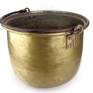 Puede incluir: Una olla de cobre antigua grande con un acabado martillado desgastado. Tiene un solo asa de hierro con remaches de cobre. La olla está aislada sobre un fondo blanco.