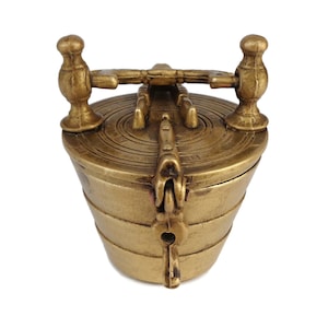 May include: A small, gold-coloured metal container with a hinged lid and two decorative handles. The container has a cylindrical shape and a textured surface.
