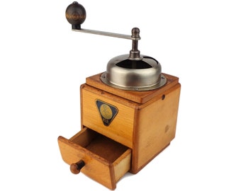 May include: A vintage wooden coffee grinder with a metal crank and a drawer for collecting the ground coffee. The grinder is made of light brown wood and has a metal top with a silver finish.