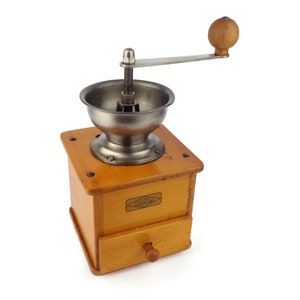 May include: A vintage wooden coffee grinder with a metal crank and a drawer. The grinder is brown and has a metal label that says "HAVA" on it.