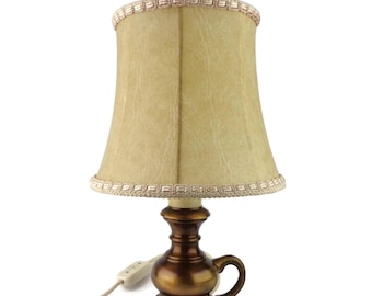 May include: A gold table lamp with a cream-colored lampshade. The lampshade has a delicate trim around the top and bottom. The lamp has a cord with a switch.