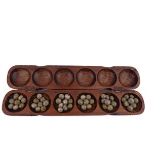 Puede incluir: Un tablero de juego de madera con 7 compartimentos, cada uno lleno de pequeñas semillas redondas verdes. El tablero está abierto y tiene un acabado de madera marrón.