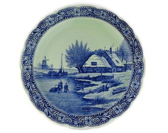 May include: A blue and white ceramic plate with a Dutch scene of a house, windmill, and figures on a canal. The plate has a decorative floral border.