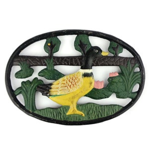 May include: Black cast iron trivet featuring a yellow and white duck design. The duck is surrounded by green leaves and pink flowers.