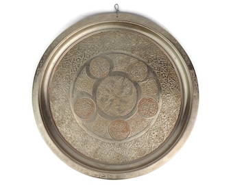 May include: A round, silver-colored metal tray with intricate engraved designs. The tray has a raised rim and a central design featuring a circle with smaller circles around it.