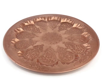 Puede incluir: Una bandeja redonda de servir de cobre con un diseño en relieve intrincado. La bandeja tiene una forma circular y un borde elevado.