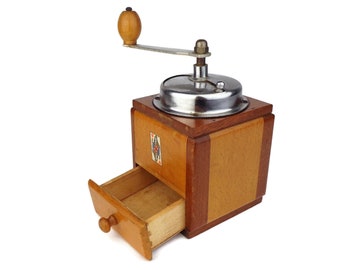May include: A vintage wooden coffee grinder with a metal grinding mechanism and a drawer to collect the ground coffee. The grinder is made of light and dark wood and has a small label on the side.