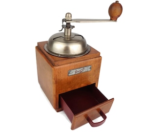 May include: A vintage wooden coffee grinder with a metal crank and a drawer. The grinder has a label that reads "Rapid".