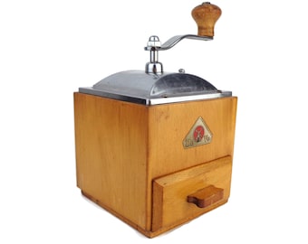 May include: A vintage wooden coffee grinder with a silver crank and a drawer for collecting the ground coffee. The grinder has a red and white label that says "De Vo" on it.
