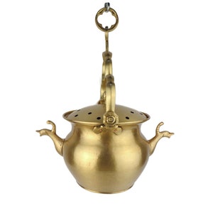Pode incluir: Um queimador de incenso de metal dourado com um corpo redondo e uma tampa com furos. O queimador tem duas alças em forma de cabeça de animal e um laço para pendurar.