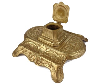 May include: A gold-toned metal inkwell with an ornate design and a hinged lid. The lid is open, revealing the empty inkwell.