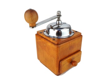 May include: A vintage wooden coffee grinder with a chrome top and a wooden crank handle. The grinder has a drawer for collecting the ground coffee.