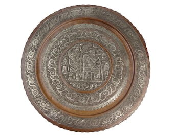 May include: A round copper tray with a silver-colored design. The design features a central image of two figures standing next to an elephant, surrounded by intricate floral patterns.