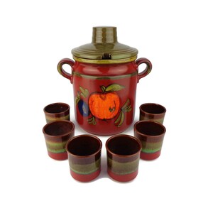 Può includere: Un'anfora in ceramica rossa con un coperchio verde e un disegno di mela dipinto. Sei tazze in ceramica rossa e verde abbinate sono disposte intorno all'anfora.