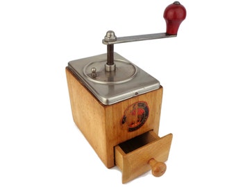 May include: A vintage wooden coffee grinder with a metal top and a drawer for storing coffee grounds. The grinder has a red handle and a label that reads "Kaffee Muhle".