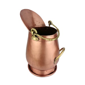 Peut inclure: Un seau en métal de couleur cuivre avec un couvercle à charnière et une poignée en laiton. Le seau a un corps arrondi et une base circulaire. La poignée est fixée au sommet du seau avec des fixations en laiton. Le couvercle est ouvert.