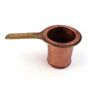 Puede incluir: Una taza medidora de color cobre con un mango de latón. La taza tiene un cuerpo cilíndrico y un borde circular con remaches. El mango es plano y rectangular. Probablemente se utiliza para medir ingredientes en la cocina.