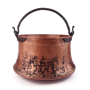 May include: A copper cauldron with a black handle and decorative features. The pot has a rounded base and a slightly flared rim. The copper has a weathered appearance with dark patches, suggesting age or use. The handle is arched and attached with ornate brackets.
